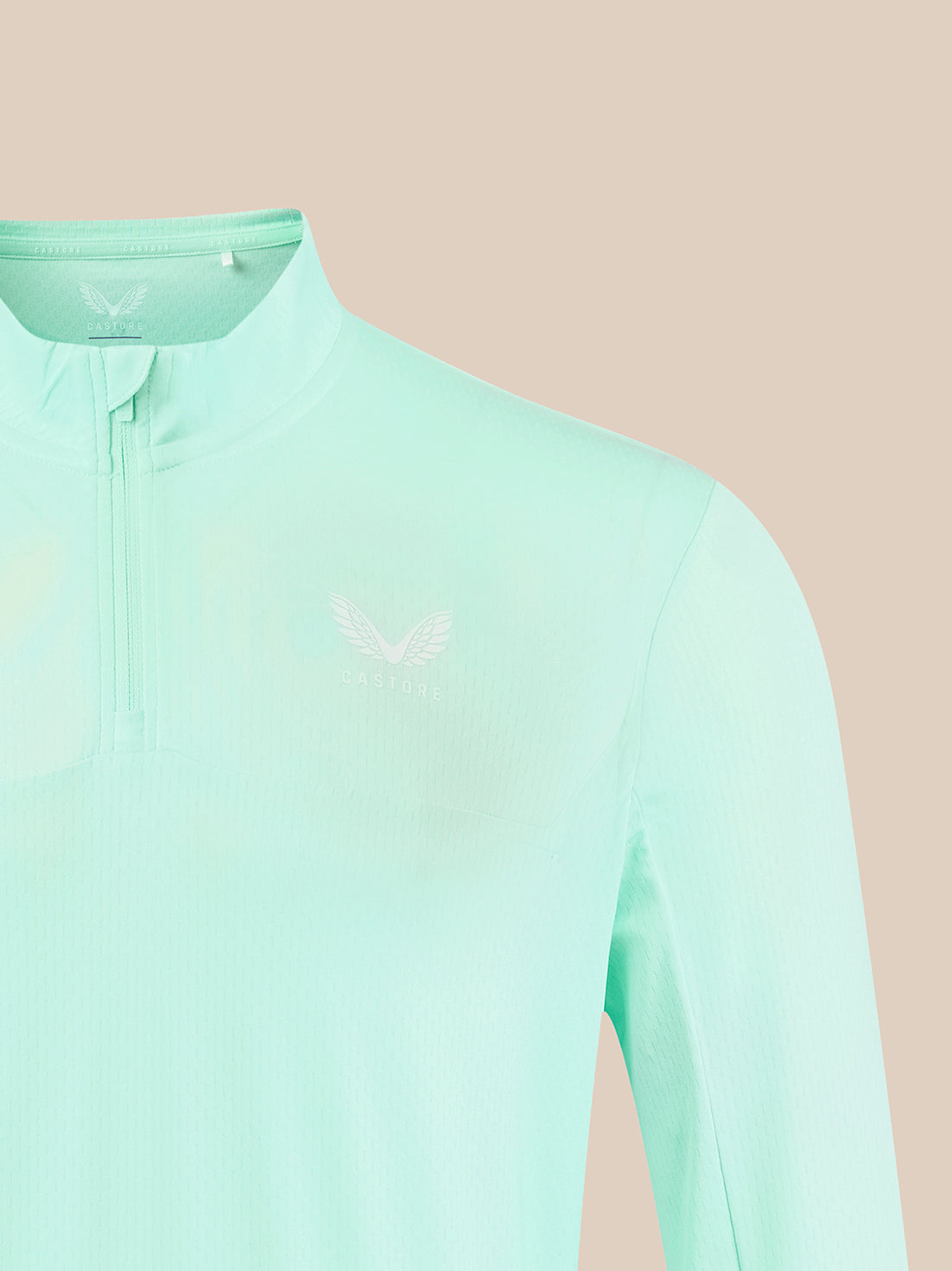 A close-up of a mint green long-sleeve athletic quarter-zip pullover with a small white wing logo on the left chest, set against a solid pale peach background.