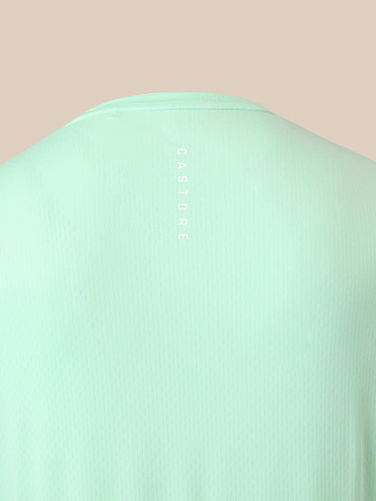Close-up of the back of a mint green ribbed t-shirt with the brand name 'CASTORE' printed vertically in white near the neckline, set against a solid pale peach background.