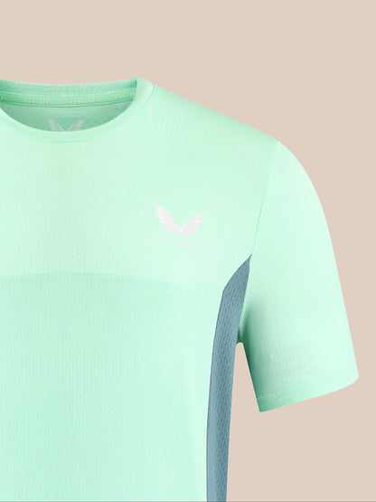 A close-up of a light mint green athletic t-shirt with a subtle textured knit pattern, featuring a small white wing logo on the left chest and a darker gray-blue mesh side panel.
