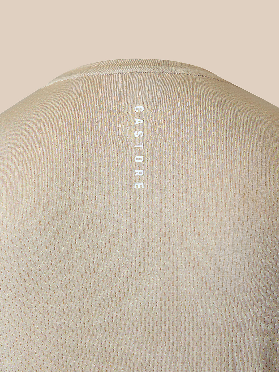 Close-up of the back of a light beige textured t-shirt with a vertical white 'CASTORE' logo printed on the center of the upper back, set against a solid beige background.