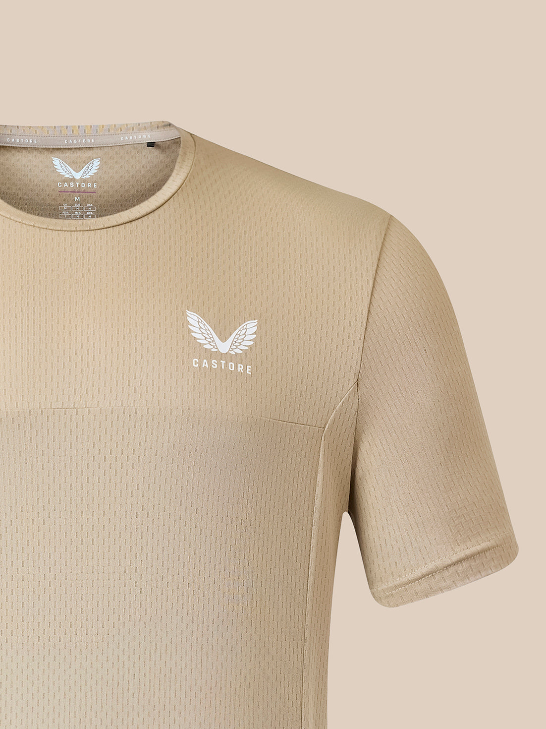 A close-up of a beige short-sleeve athletic shirt with a textured waffle knit pattern, featuring a white winged logo and the brand name 'CASTORE' on the left chest.