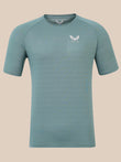 A short-sleeve athletic t-shirt in a muted teal-blue color, featuring a white winged logo and the brand name 'Castore' on the left chest, displayed against a solid beige background.