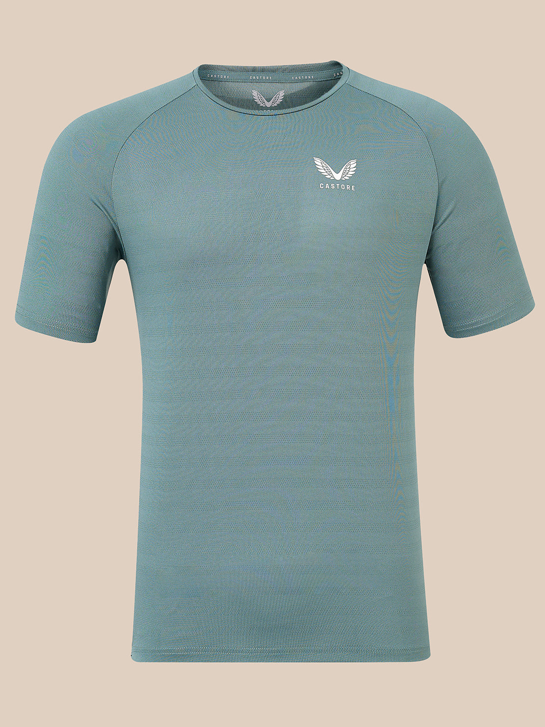 A short-sleeve athletic t-shirt in a muted teal-blue color, featuring a white winged logo and the brand name 'Castore' on the left chest, displayed against a solid beige background.