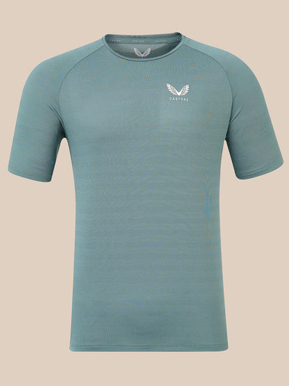 A short-sleeve athletic t-shirt in a muted teal-blue color, featuring a white winged logo and the brand name 'Castore' on the left chest, displayed against a solid beige background.