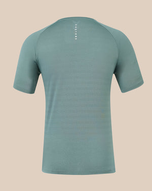 A short-sleeve athletic t-shirt shown from the back, featuring a textured knit fabric in a muted sage green color with a vertical white 'Castore' logo printed on the upper back, set against a solid beige background.