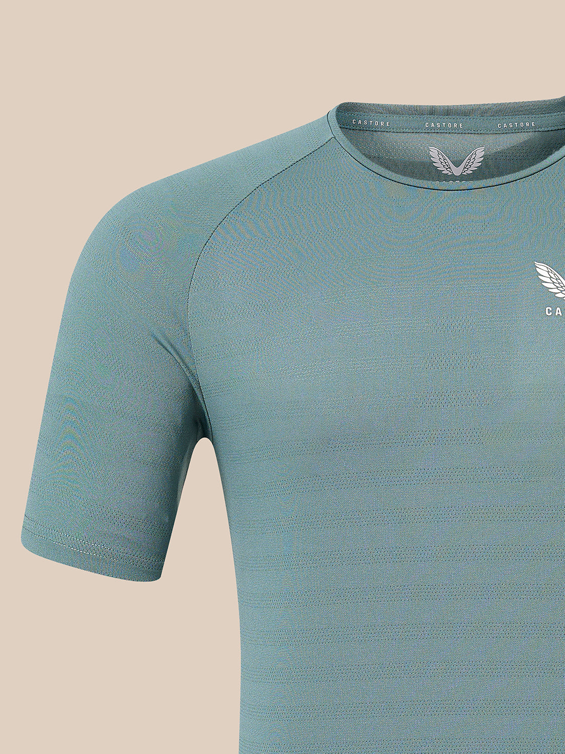A close-up of a short-sleeve athletic t-shirt in a muted teal-blue color, featuring a textured knit fabric, a winged logo on the chest, and a matching inner neck label, set against a solid beige background.