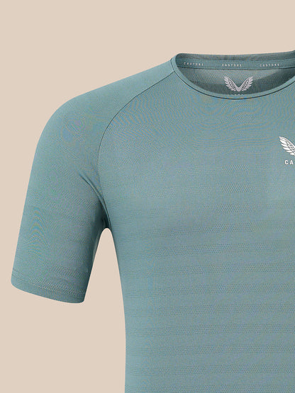 A close-up of a short-sleeve athletic t-shirt in a muted teal-blue color, featuring a textured knit fabric, a winged logo on the chest, and a matching inner neck label, set against a solid beige background.