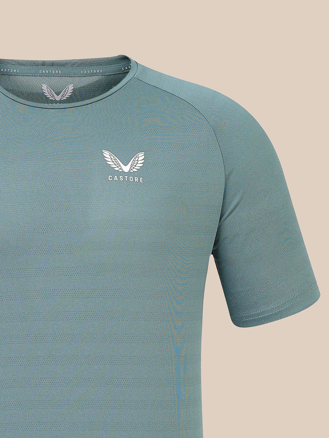 A close-up of a short-sleeve athletic t-shirt in a muted teal-blue color, featuring a white winged logo and the brand name 'CASTORE' on the left chest, set against a solid beige background.