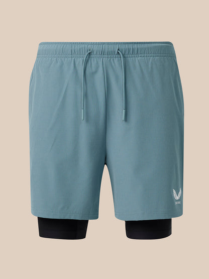 A pair of light blue athletic shorts with a matching elastic drawstring waist, featuring a black compression liner visible at the hem and a small white wing logo on the lower right leg, set against a solid beige background.
