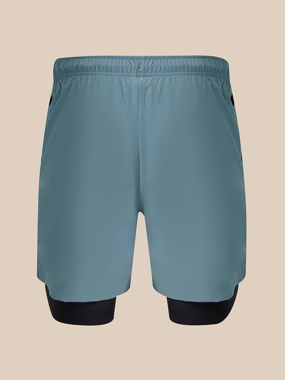 A pair of athletic shorts in a muted slate blue color, featuring a built-in black compression layer visible at the lower legs, shown against a solid beige background.