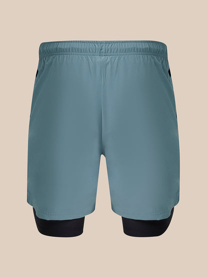 A pair of athletic shorts in a muted slate blue color, featuring a built-in black compression layer visible at the lower legs, shown against a solid beige background.