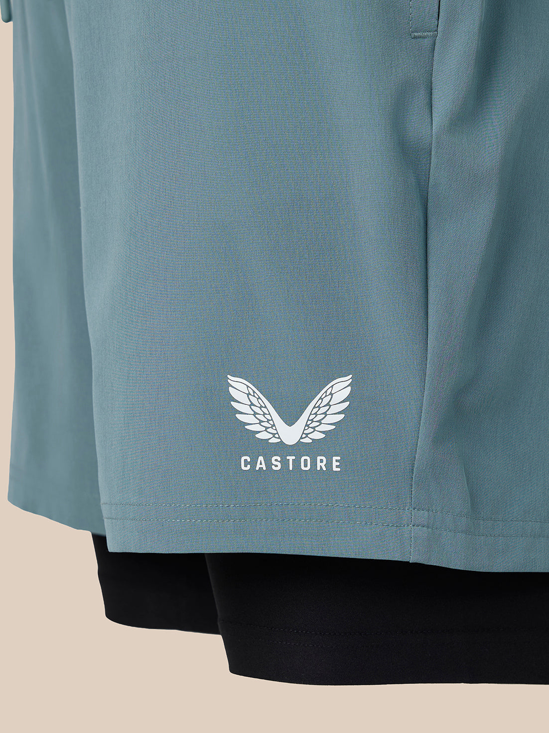 Close-up of a dusty blue athletic top with a white Castore wing logo and brand name printed on the front, layered over black leggings against a light beige background.
