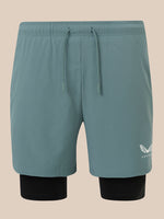 A pair of men's athletic shorts in a muted teal-blue color, featuring a matching drawstring waist and a black inner layer visible at the legs. A small white embroidered wing logo with 'CASTORE' text is on the lower left leg.