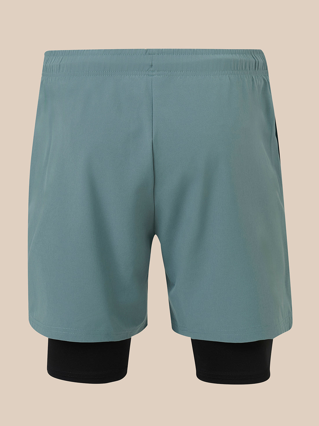 A pair of athletic shorts in a muted teal-blue color, featuring a built-in black compression liner visible at the lower legs, shown against a solid beige background.