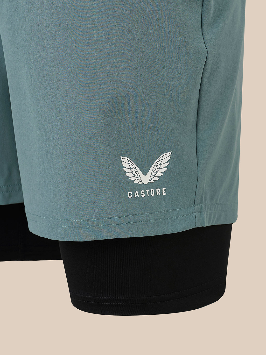 Close-up of teal athletic shorts with a white Castore wing logo and text on the lower left leg, worn over black leggings against a light beige background.