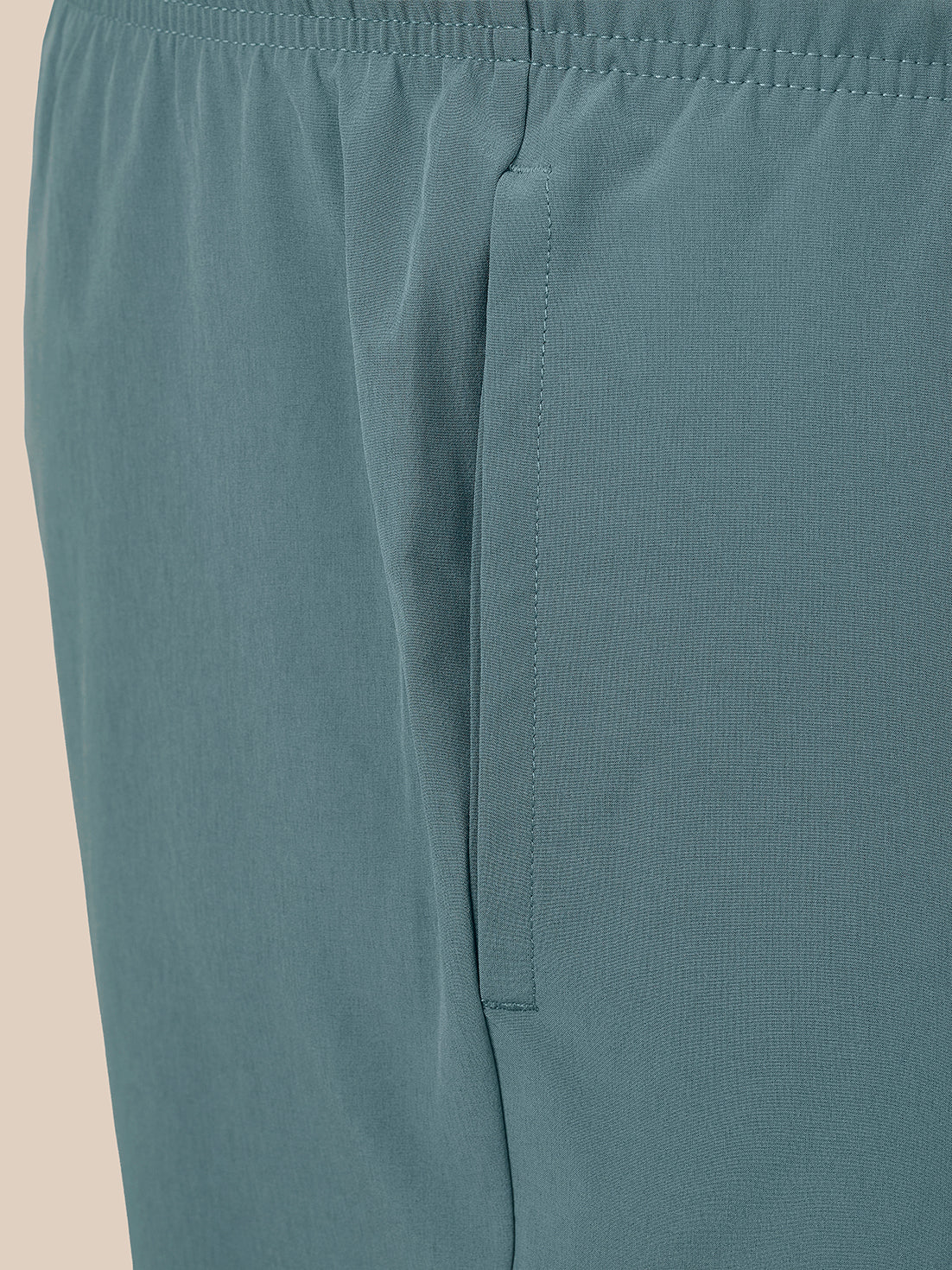 Close-up of a pair of muted teal athletic shorts showing the waistband, side seam, and a vertical welt pocket.