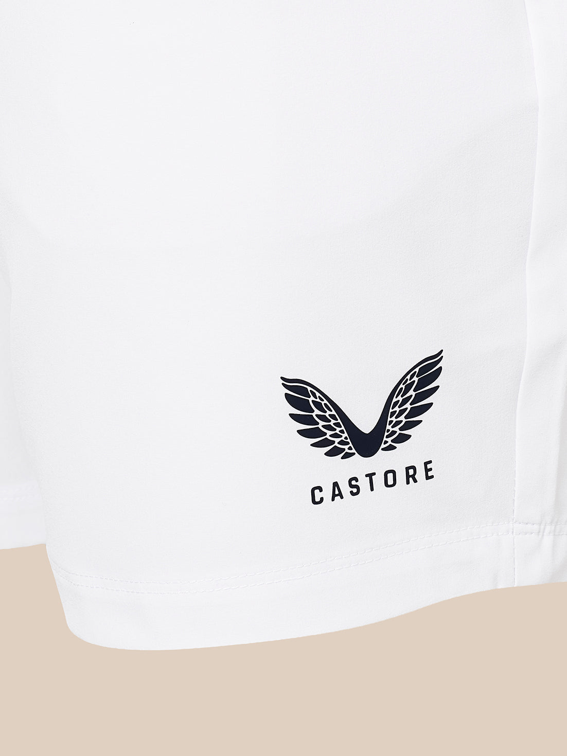 Close-up of a white garment, likely shorts or a skirt, featuring a black embroidered winged logo and the brand name 'CASTORE' printed below it. The fabric is shown against a neutral beige background.