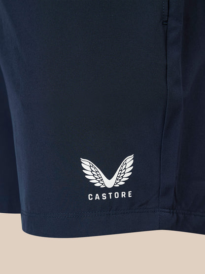 Close-up of navy athletic shorts with a white Castore wing logo and brand name printed on the lower left leg, set against a light beige background.
