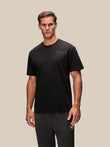 A man modeling a black short-sleeve crewneck t-shirt with a small white 'CASTORE' logo on the left chest, paired with dark gray athletic pants, against a solid beige background.
