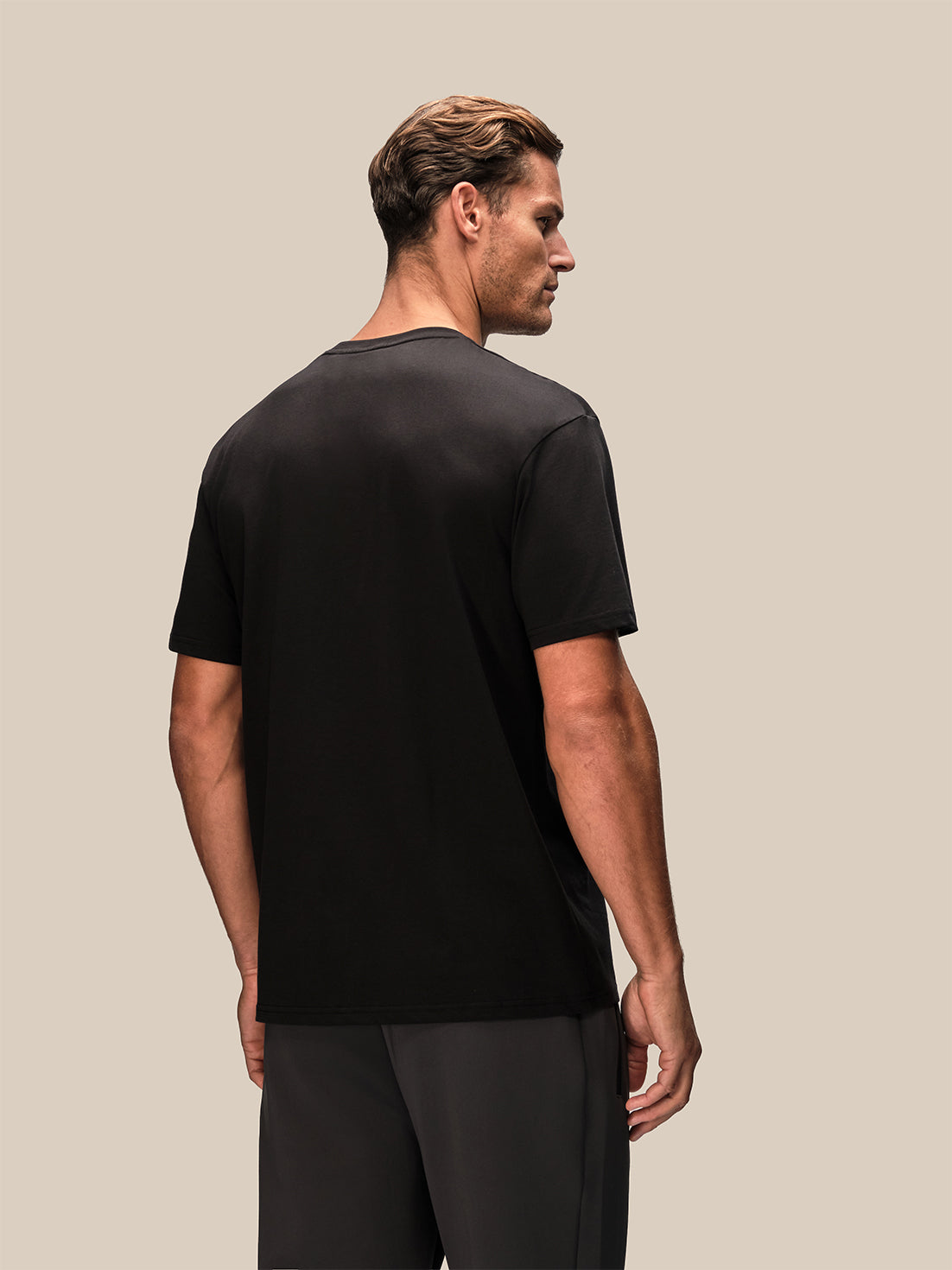 A man shown from the back wearing a black short-sleeve crewneck t-shirt and dark gray athletic pants, set against a solid light beige background.
