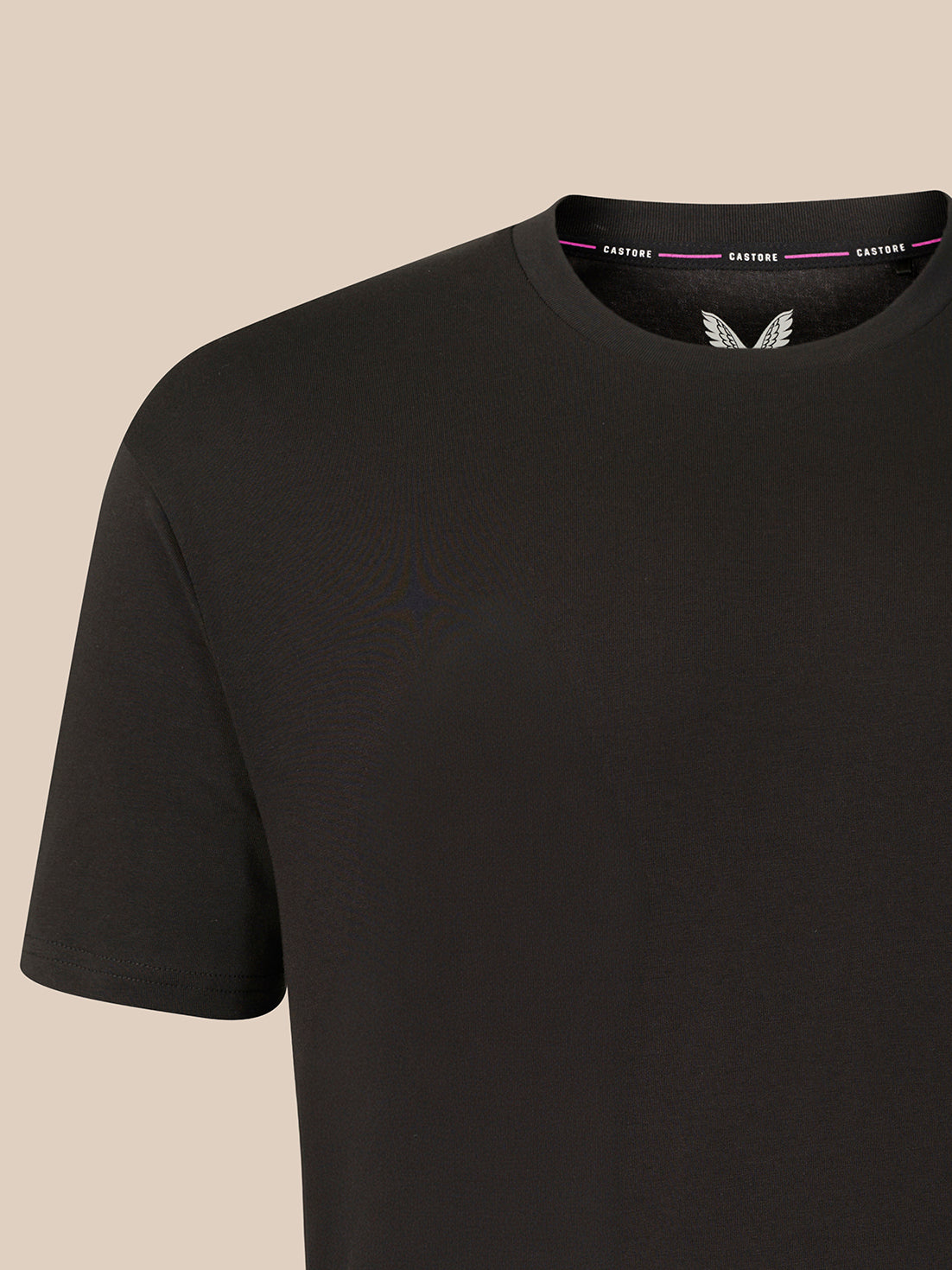A close-up of a black short-sleeve crewneck t-shirt on a light beige background, showing a white wing logo on the inside of the collar and the brand name 'CASTORE' printed on the inner neckband.
