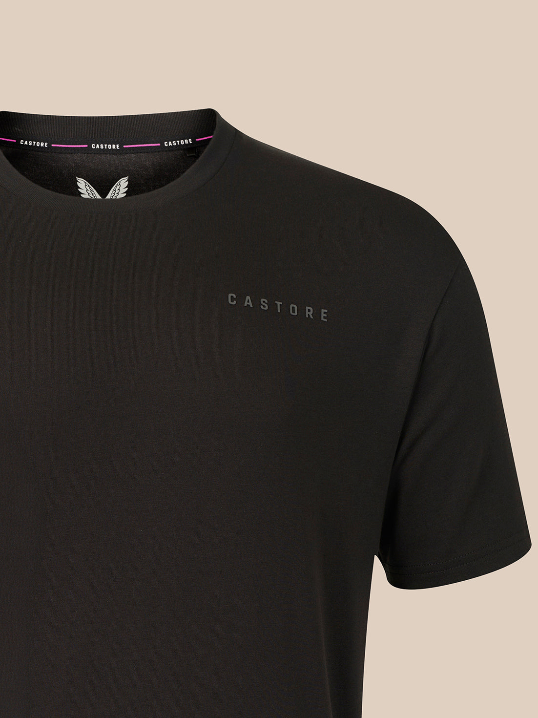 A close-up of a black Castore short-sleeve t-shirt with a subtle tonal logo on the left chest, set against a solid beige background.
