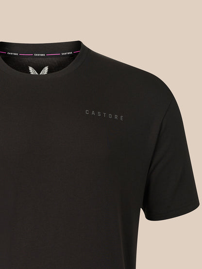 A close-up of a black Castore short-sleeve t-shirt with a subtle tonal logo on the left chest, set against a solid beige background.