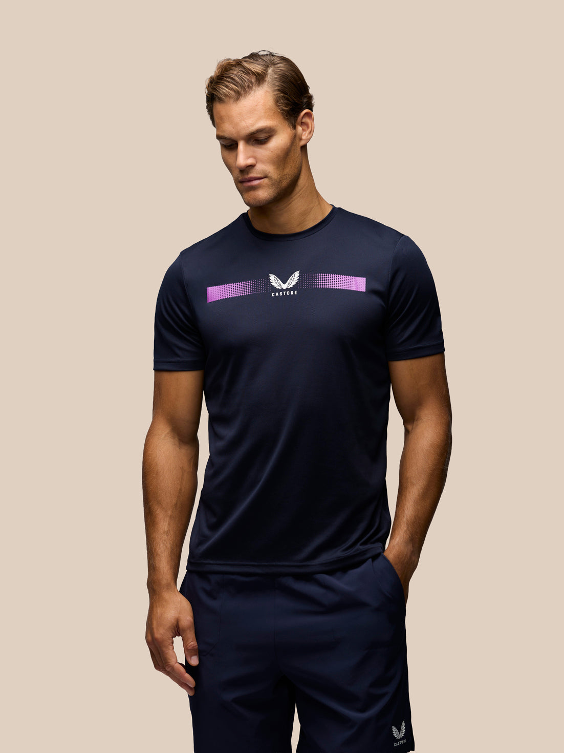 A man wearing a navy blue athletic t-shirt and matching shorts with a purple and white chest graphic, posed against a solid beige background.