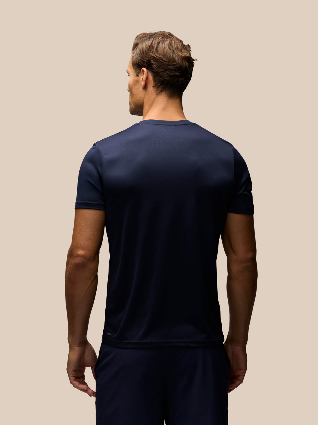 A man viewed from behind wearing a dark navy blue short-sleeve athletic t-shirt and matching navy shorts, set against a solid beige background.