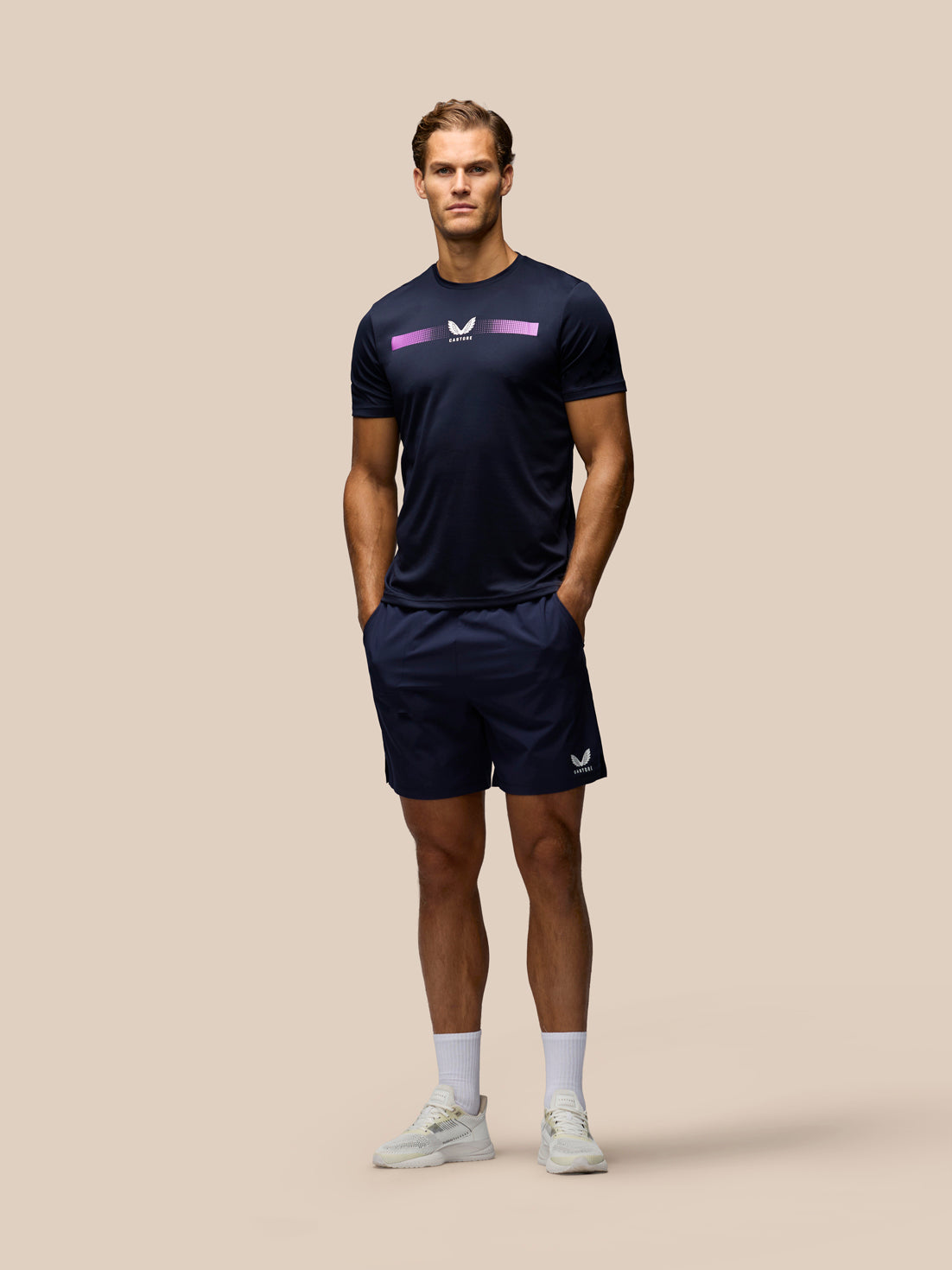 A male model stands against a solid beige background wearing a navy athletic t-shirt with a purple and white logo, matching navy athletic shorts, white socks, and white sneakers.