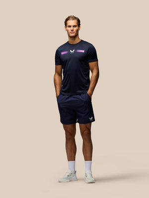 A male model stands against a solid beige background wearing a navy athletic t-shirt with a purple and white logo, matching navy athletic shorts, white socks, and white sneakers.