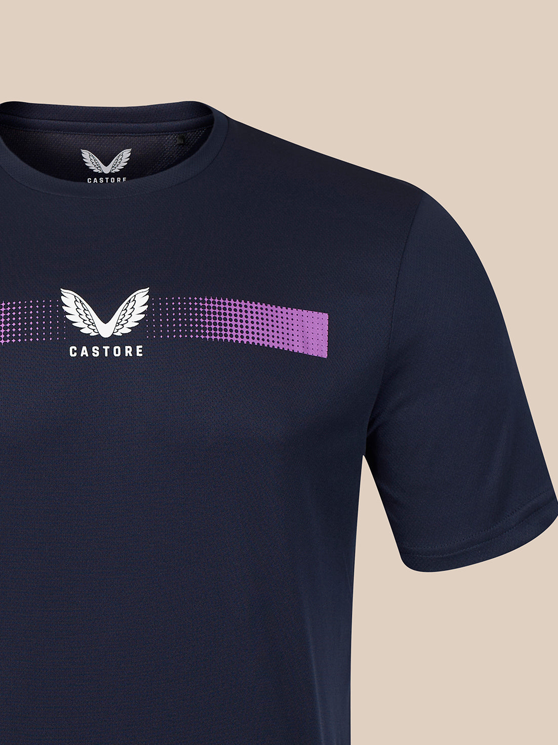 A close-up of a navy blue Castore short-sleeve t-shirt featuring a white winged logo and a horizontal purple and white dotted graphic on the chest, set against a solid beige background.