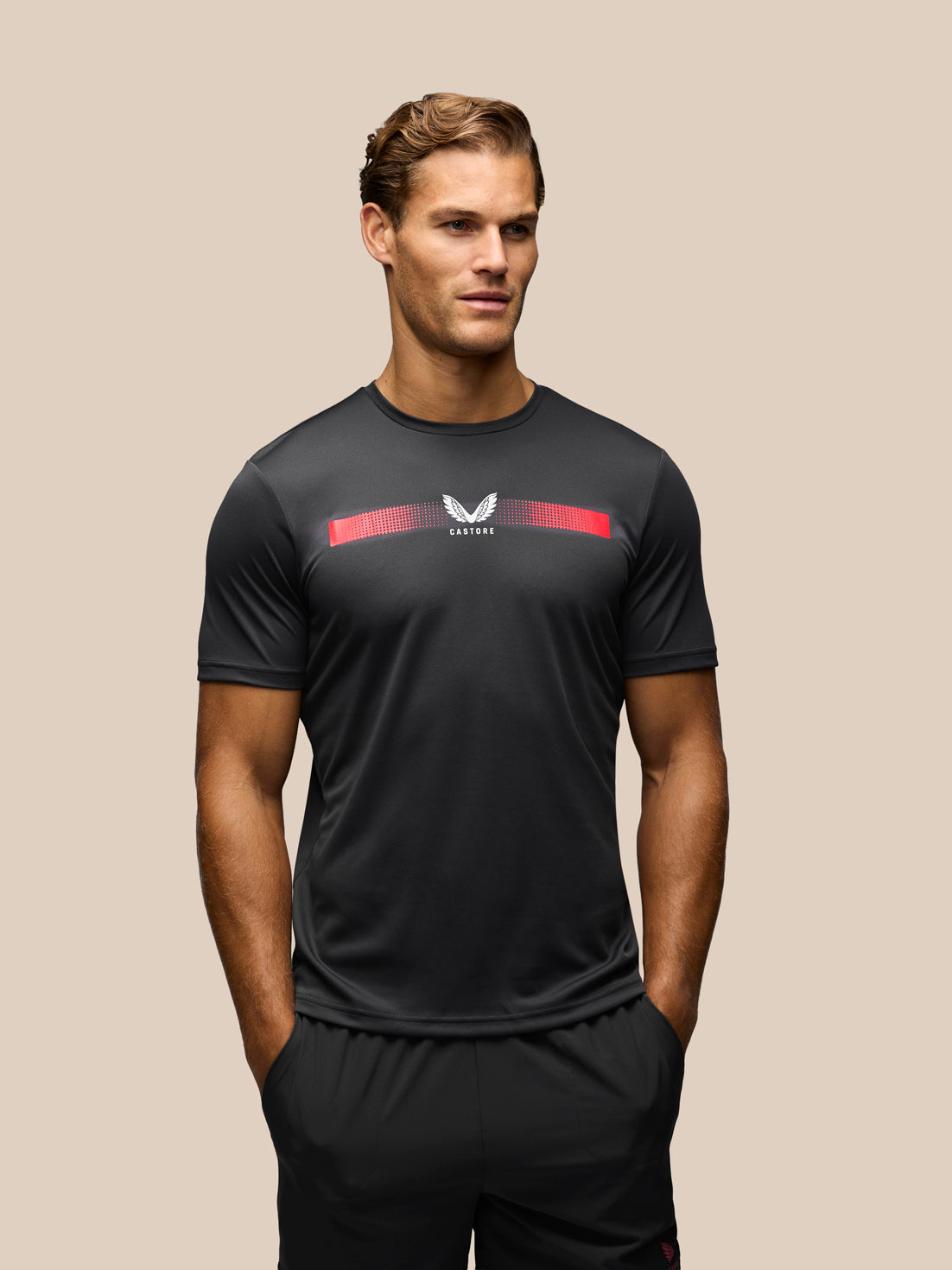 A man wearing a black athletic t-shirt with a red and white chest graphic, paired with black shorts, set against a solid beige background.