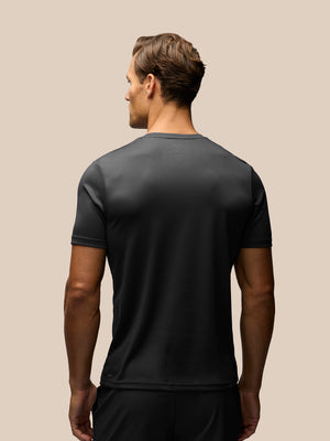 A man viewed from the back wearing a dark charcoal gray athletic t-shirt and black shorts, set against a solid beige background.
