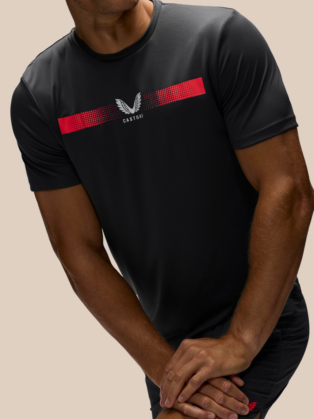 A man wearing a black short-sleeve athletic t-shirt with a red chest stripe and a white winged logo, shown from the neck down against a solid beige background.