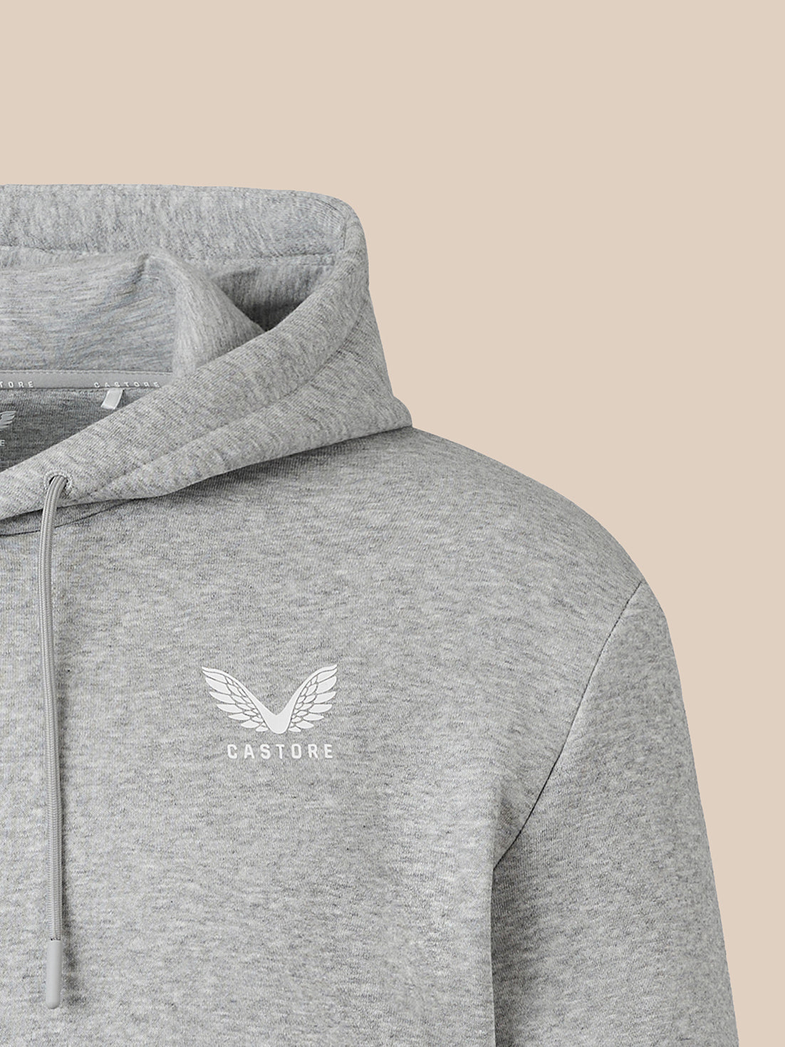 Close-up of a heather gray pullover hoodie with a white winged logo and the word 'CASTORE' on the left chest, shown against a plain beige background.