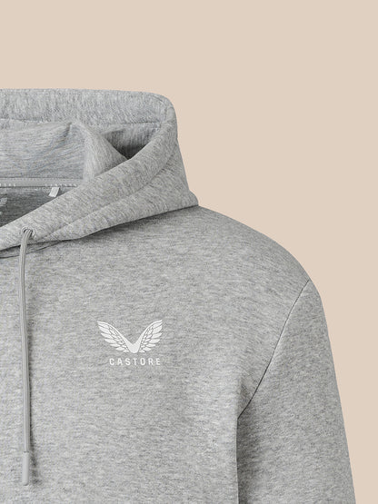 Close-up of a heather gray pullover hoodie with a white winged logo and the word 'CASTORE' on the left chest, shown against a plain beige background.