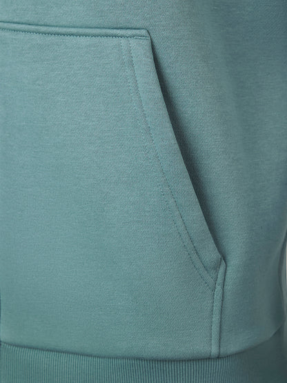 Close-up of a dusty teal-green hoodie showing the texture of the fabric and the edge of the front kangaroo pocket.