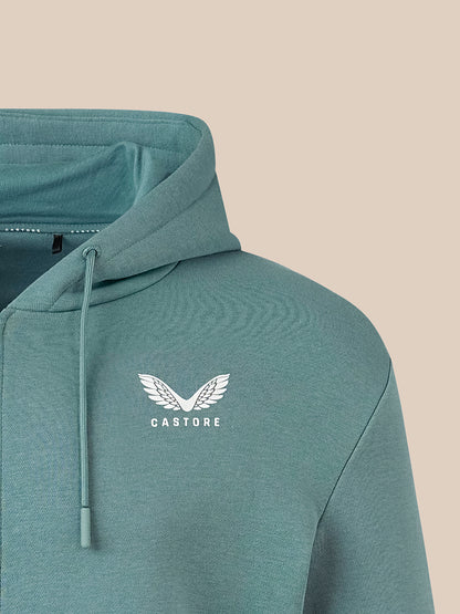 A close-up of a teal-green hooded sweatshirt with a white winged logo and the word 'CASTORE' on the chest, set against a solid beige background.