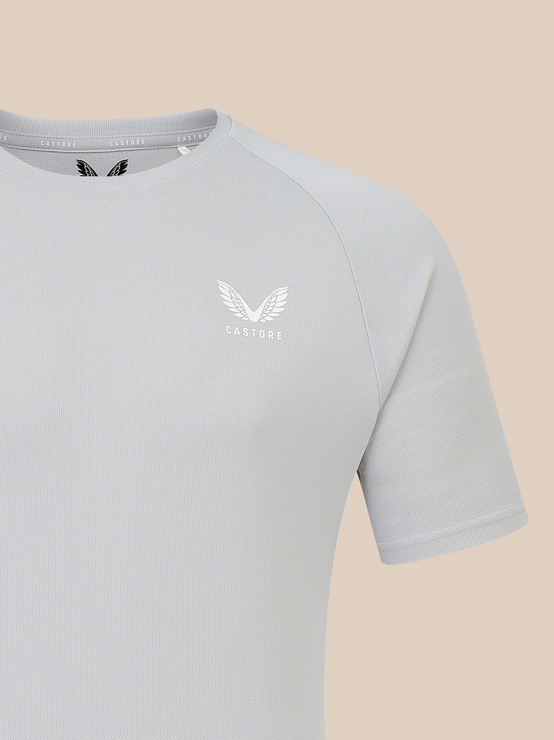 A close-up of a light gray short-sleeve athletic t-shirt with a white winged logo and the brand name 'CASTORE' printed on the left chest, set against a solid beige background.