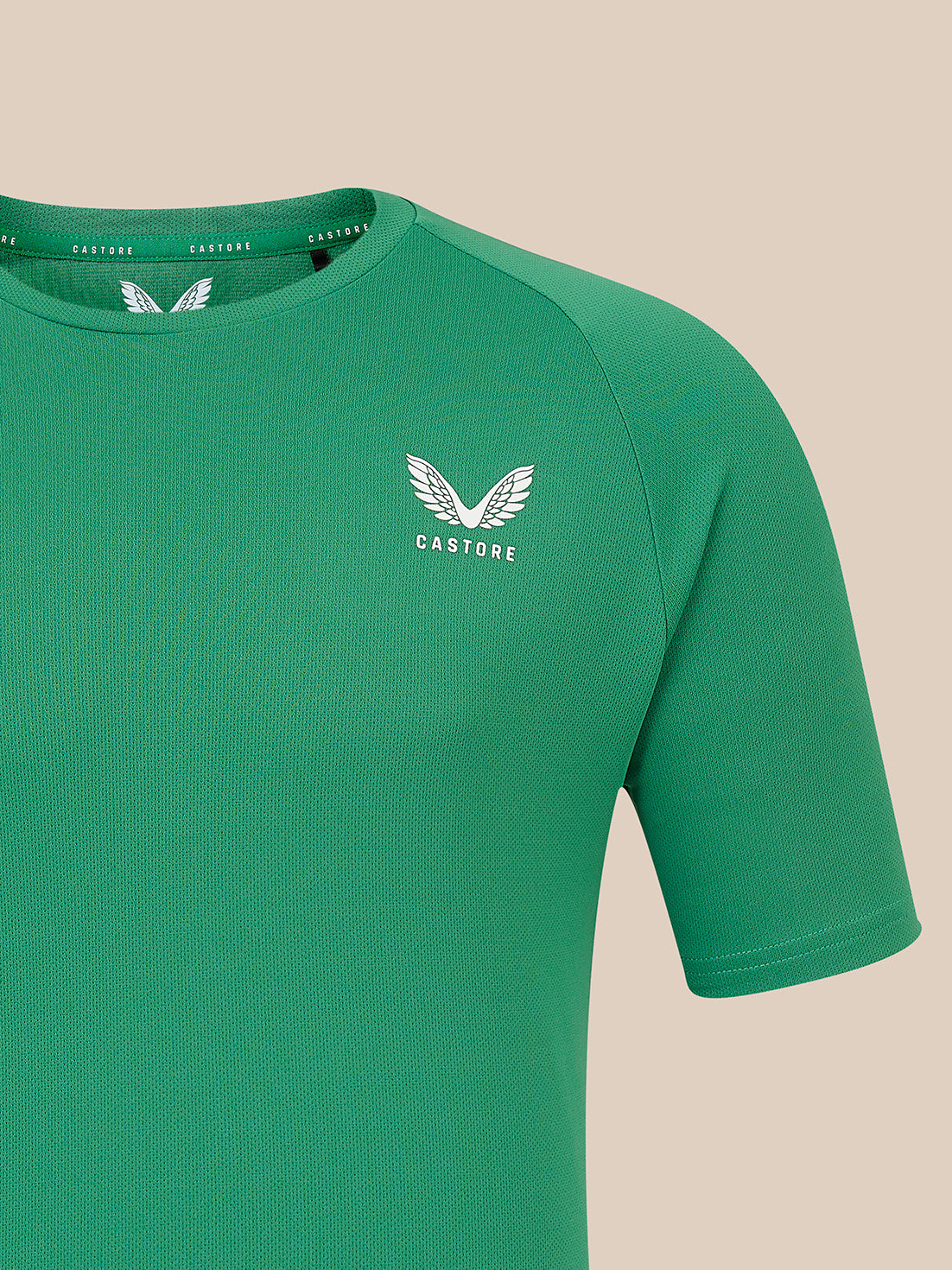 A close-up of a green short-sleeve athletic t-shirt with a white winged logo and the brand name 'CASTORE' printed on the left chest, set against a solid beige background.