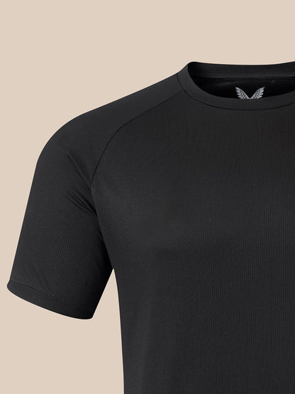 Close-up of a black short-sleeve athletic t-shirt with raglan sleeves and a small silver wing logo on the inside of the collar, set against a solid beige background.