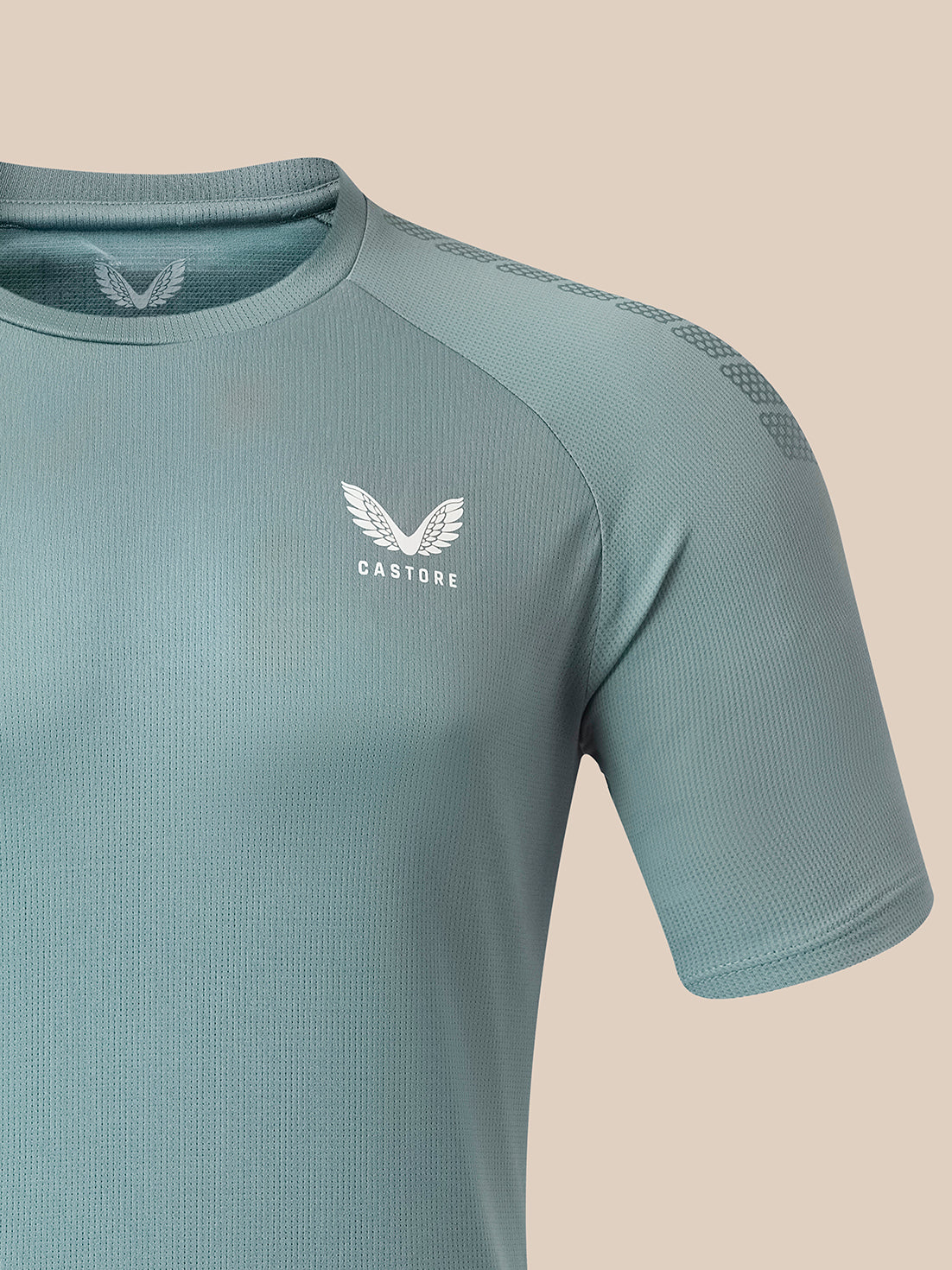 A close-up of a short-sleeve athletic shirt in a muted sage green color, featuring a white winged logo and the brand name 'CASTORE' on the left chest, set against a solid beige background.