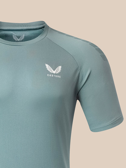 A close-up of a short-sleeve athletic shirt in a muted sage green color, featuring a white winged logo and the brand name 'CASTORE' on the left chest, set against a solid beige background.