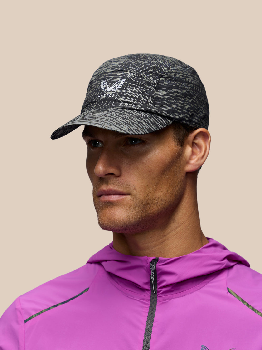 A man wearing a dark gray heathered athletic cap with a white wing logo and the brand name CASTORE' on the front, paired with a bright pink athletic jacket, set against a solid beige background.