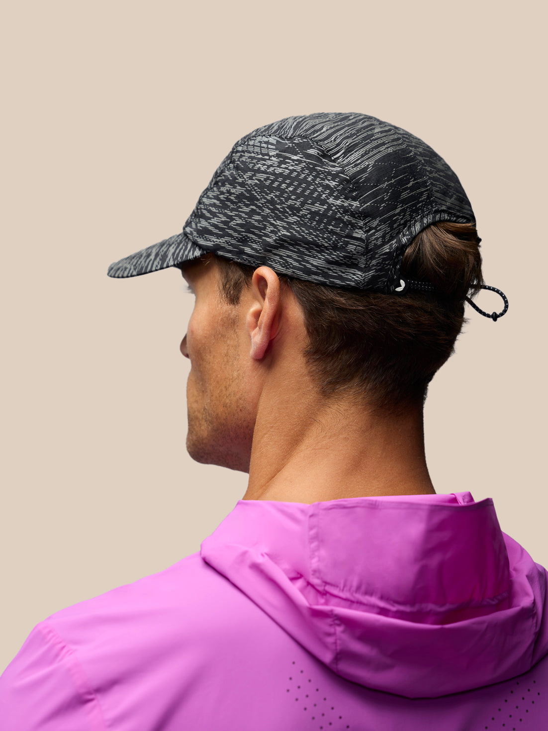 A man shown from the back wearing a dark gray and black patterned athletic cap and a bright pink hooded jacket, against a solid beige background.