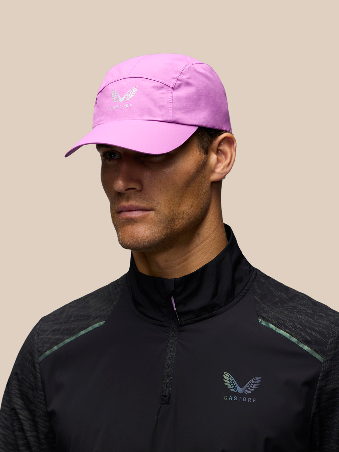 A man wearing a light pink running cap with a white wing logo and a black athletic quarter-zip jacket with mint green accents, set against a solid beige background.