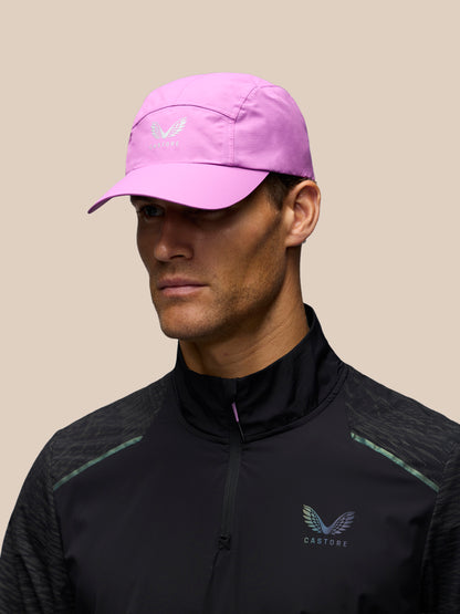 A man wearing a light pink running cap with a white wing logo and a black athletic quarter-zip jacket with mint green accents, set against a solid beige background.