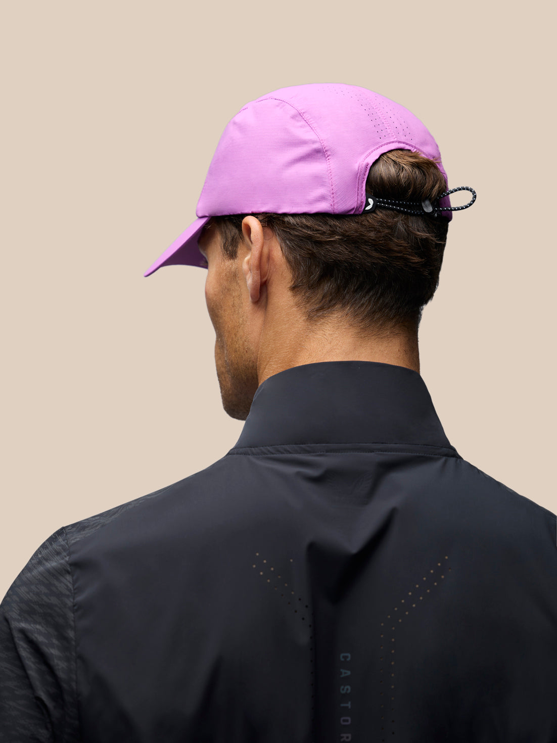 A man shown from behind wearing a light purple baseball cap and a black performance jacket against a solid beige background.