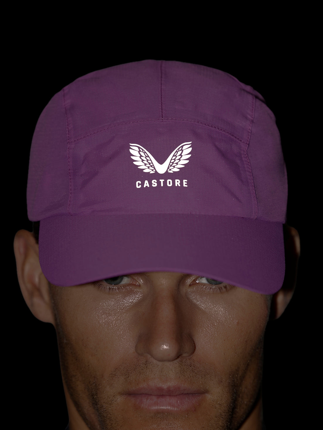 A man wearing a purple baseball cap with a white embroidered wing logo and the brand name 'CASTORE' on the front, set against a solid black background.